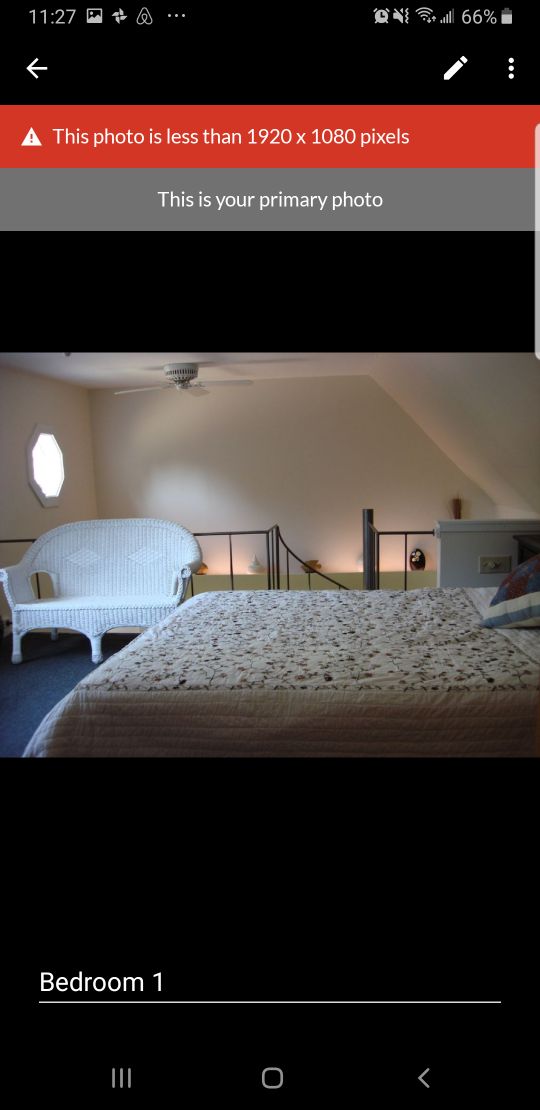 The bedroom features a queen bed covered with a floral patterned quilt. A cozy white wicker chair is positioned beside the bed. A unique octagonal window allows natural light to enter, while a fan is mounted on the ceiling for comfort.