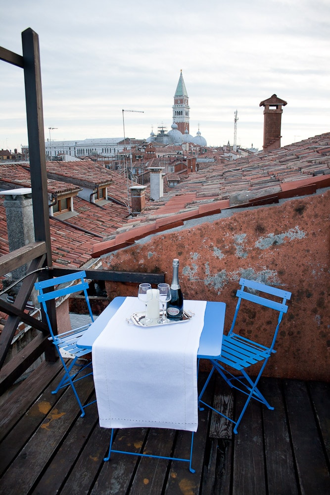 The private rooftop terrace features a small blue table and two matching chairs, set for dining. A bottle of champagne and glasses are arranged on a tablecloth, with a view of the city's rooftops and bell tower in the background under an overcast sky.