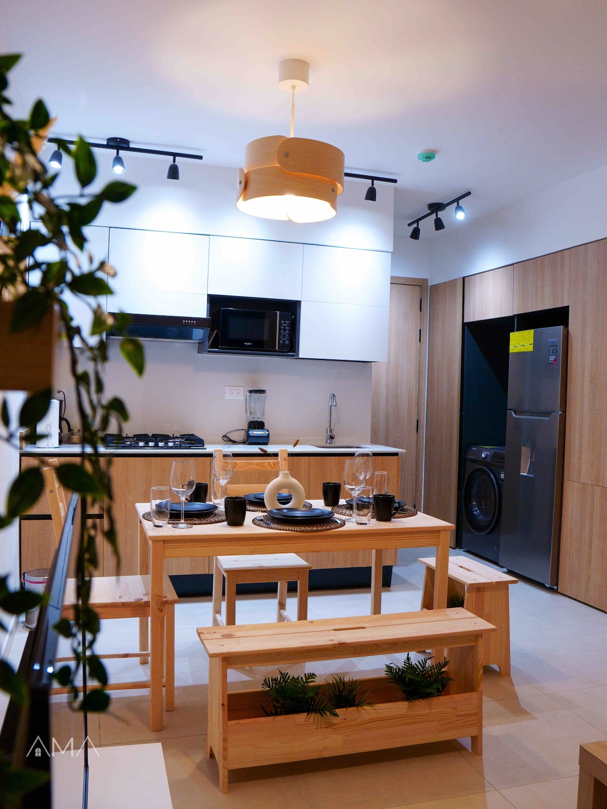 A modern kitchen area features a wooden dining table set for four, accompanied by benches and stylish dinnerware. A sleek light fixture hangs above, while cabinetry and appliances, including a microwave and refrigerator, are neatly arranged. An indoor plant adds a touch of greenery to the space.