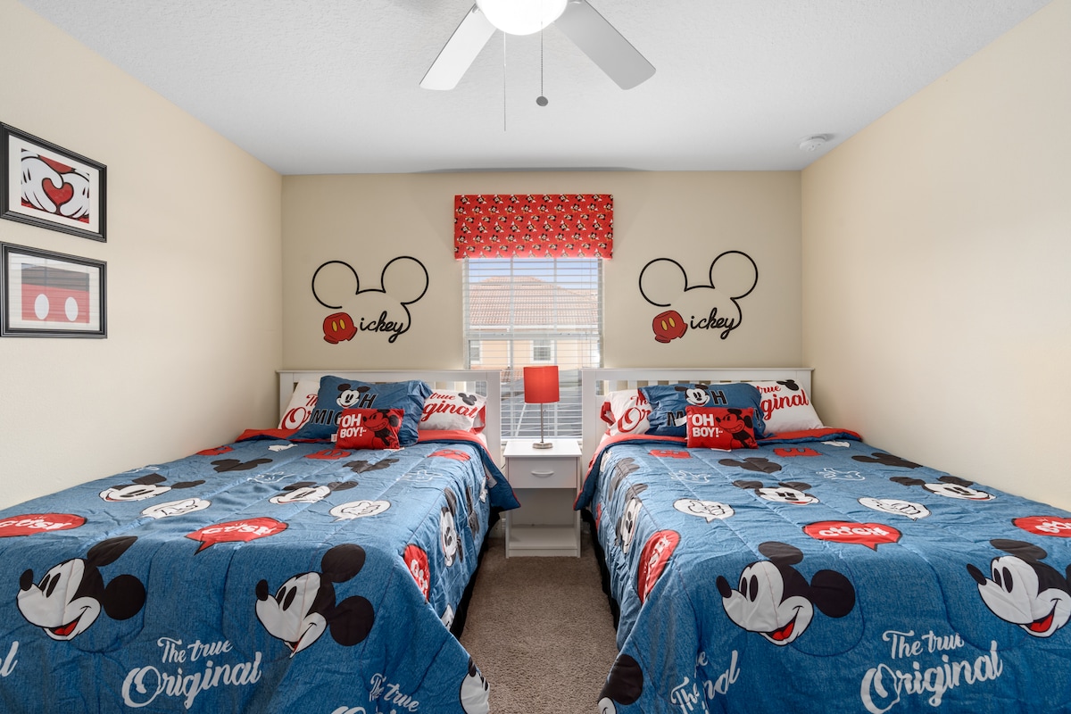 A cheerful bedroom featuring two spacious beds adorned with vibrant Mickey Mouse-themed bedding. Two playful Mickey Mouse wall decals are displayed above each bed, along with a decorative window treatment in coordinating colors. A ceiling fan provides airflow in the room with ample natural light from the window.