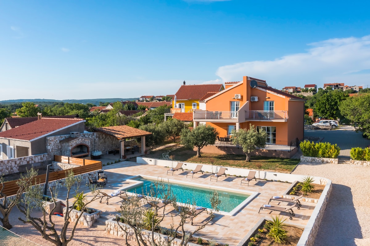 An aerial view captures a spacious villa with an orange exterior, surrounded by lush greenery and distant rooftops. Below, a serene swimming pool is framed by sun loungers and garden beds featuring olive trees and shrubs, offering a calm and inviting outdoor space.