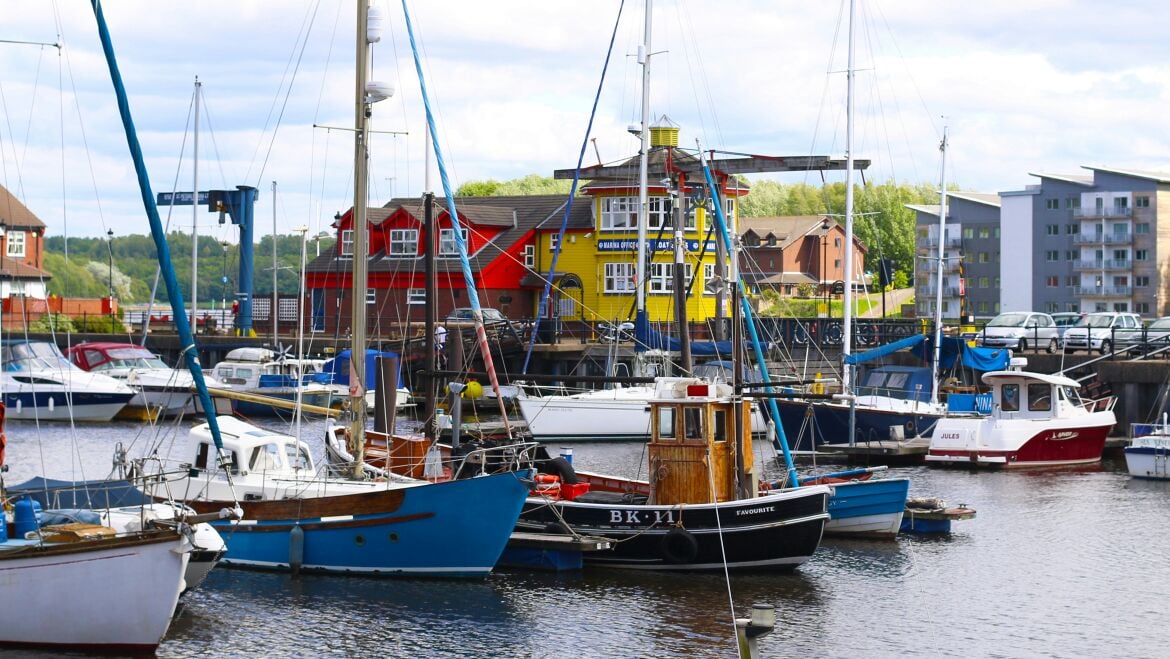 A marina scene is presented, featuring a variety of boats moored in calm waters. Colorful buildings, including a bright yellow structure, form the backdrop, while masts and rigging add vertical lines to the composition.