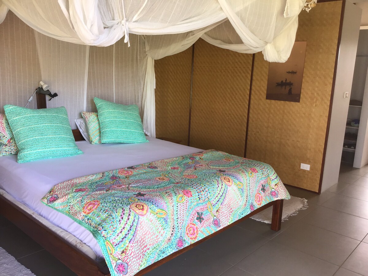 A spacious bed is draped with a light canopy, featuring vibrant decorative pillows. The bedspread showcases intricate patterns in soft pastel colors. A woven screen adds a touch of texture to the background, while a doorway hints at adjacent spaces in the pavilion.