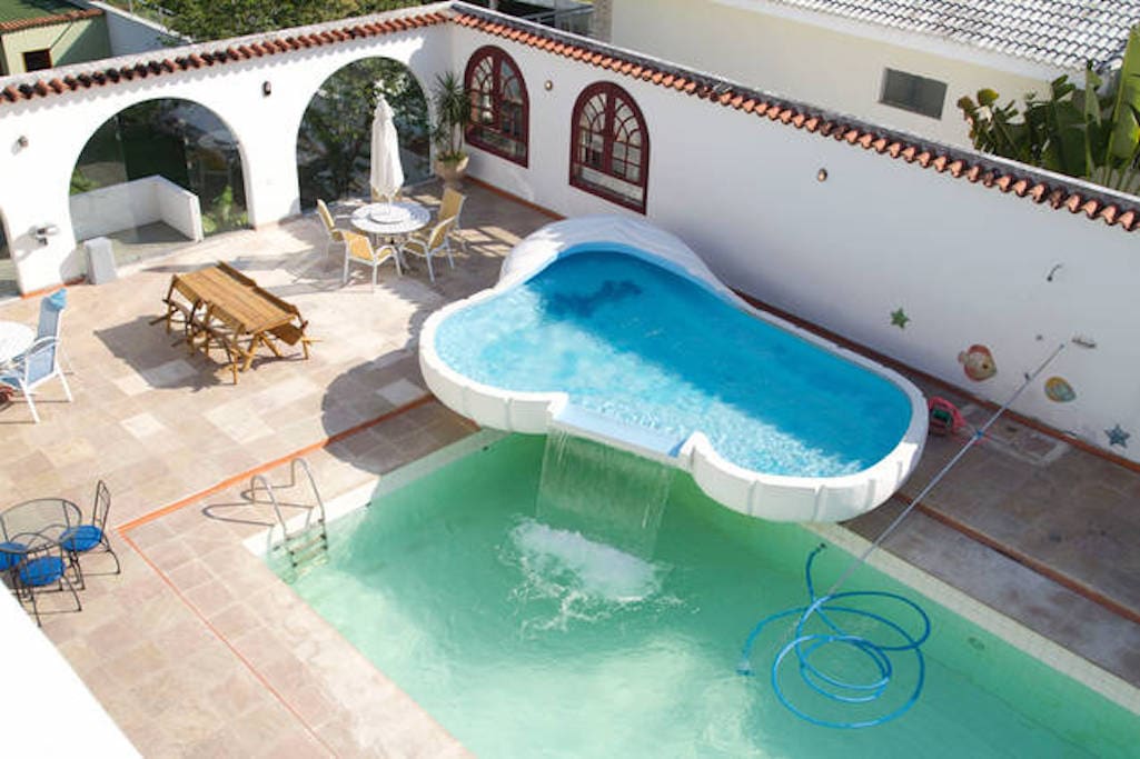 An outdoor area features a unique pool with a waterfall, complemented by poolside lounging spaces. Tables and chairs are arranged around the pool, with a sun umbrella providing shade. The surrounding walls are adorned with decorative elements, and greenery enhances the environment.