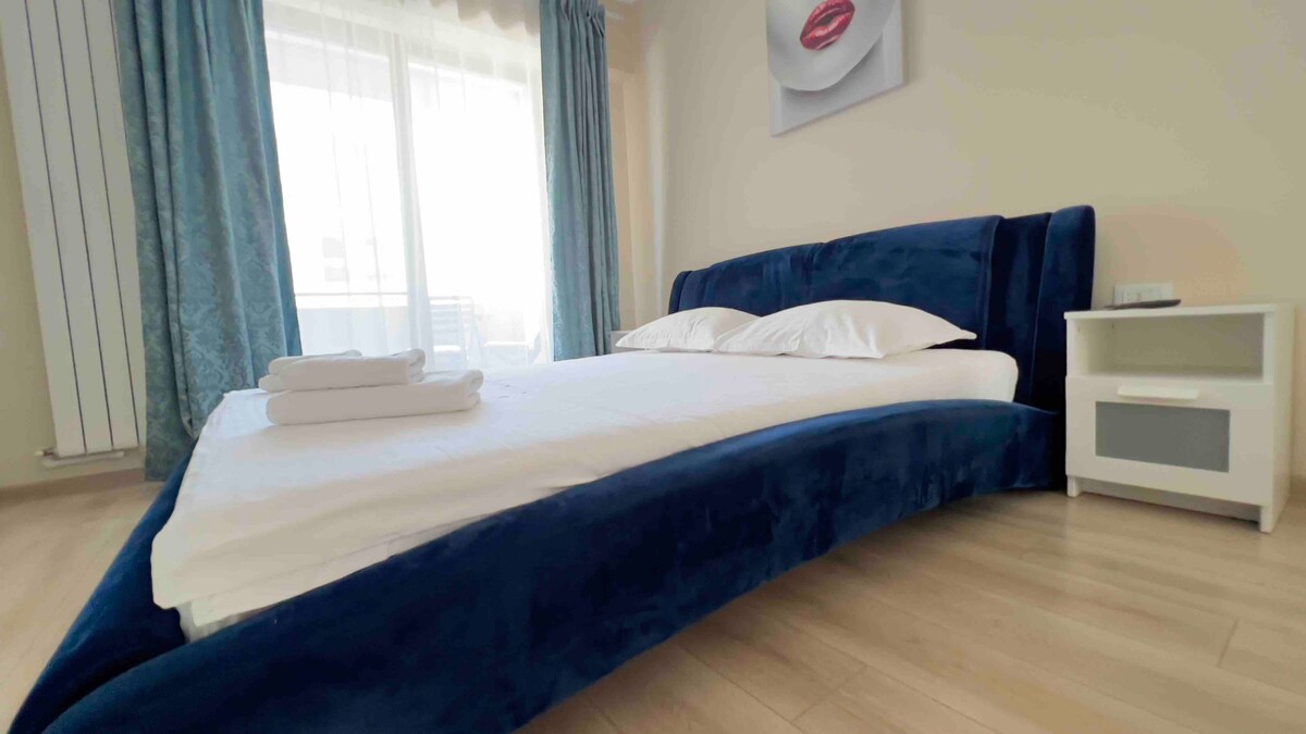 A spacious bedroom features a modern velvet bed with soft white linens. Natural light enters through large windows, adorned with elegant curtains, while a bedside table stands nearby, complementing the room's contemporary design. A piece of wall art adds a touch of character.