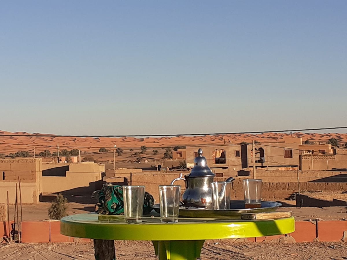 A circular green table is set outdoors, featuring a traditional metal teapot and four clear glasses. The background reveals the undulating dunes of a desert landscape under a clear sky, with simple buildings made of earth tones nestled in the distance.