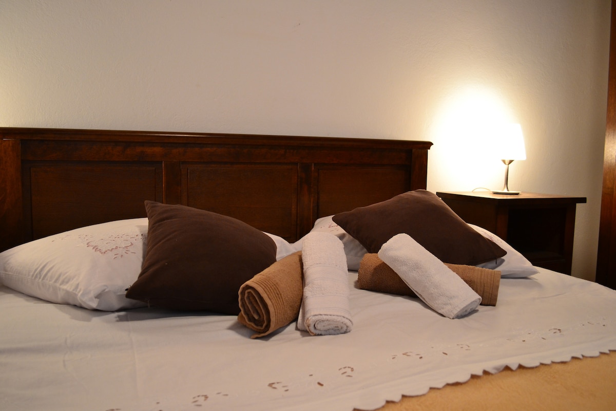 A spacious bed is neatly arranged with multiple pillows and neatly rolled towels. A bedside lamp softly illuminates the scene, casting a warm glow against the wooden headboard and the serene bedding.