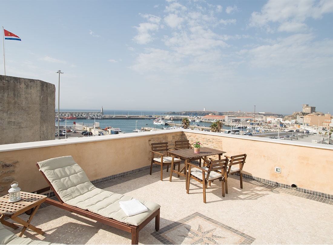 A rooftop terrace offers expansive views of the harbor and surrounding area. A lounge chair and a set of wooden dining furniture are present, providing a comfortable space to relax. The sky is bright and partly cloudy, enhancing the serene atmosphere.