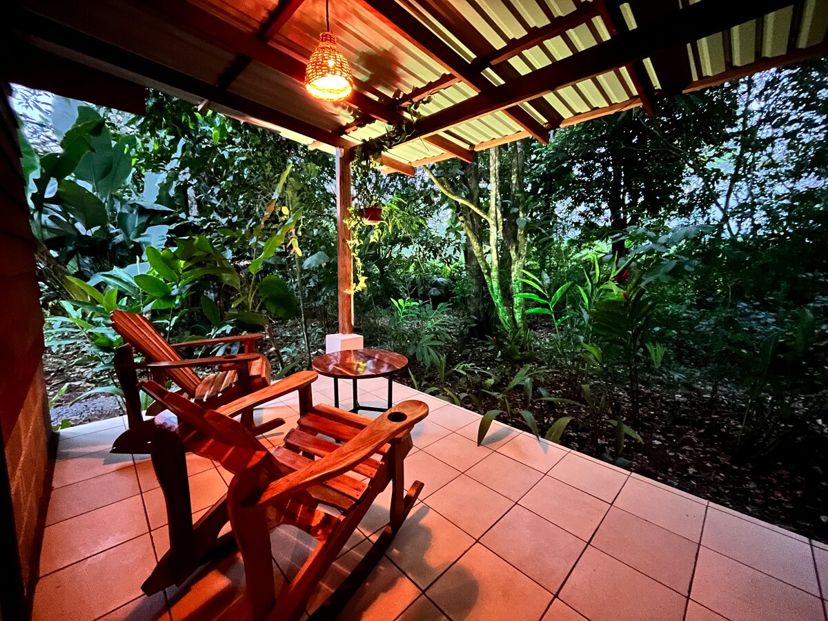 A shaded terrace is furnished with two wooden chairs and a small table, surrounded by lush greenery. Soft lighting creates a relaxing ambiance, highlighting the vibrant plants that border the area. This serene outdoor space invites guests to unwind in nature.