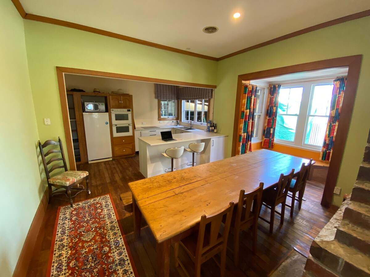 A spacious kitchen area features a long wooden dining table with chairs, complemented by a vibrant area rug. A modern kitchen counter with bar stools overlooks the cooking space, equipped with a refrigerator and an oven. Large windows framed by colorful curtains allow natural light to fill the room.