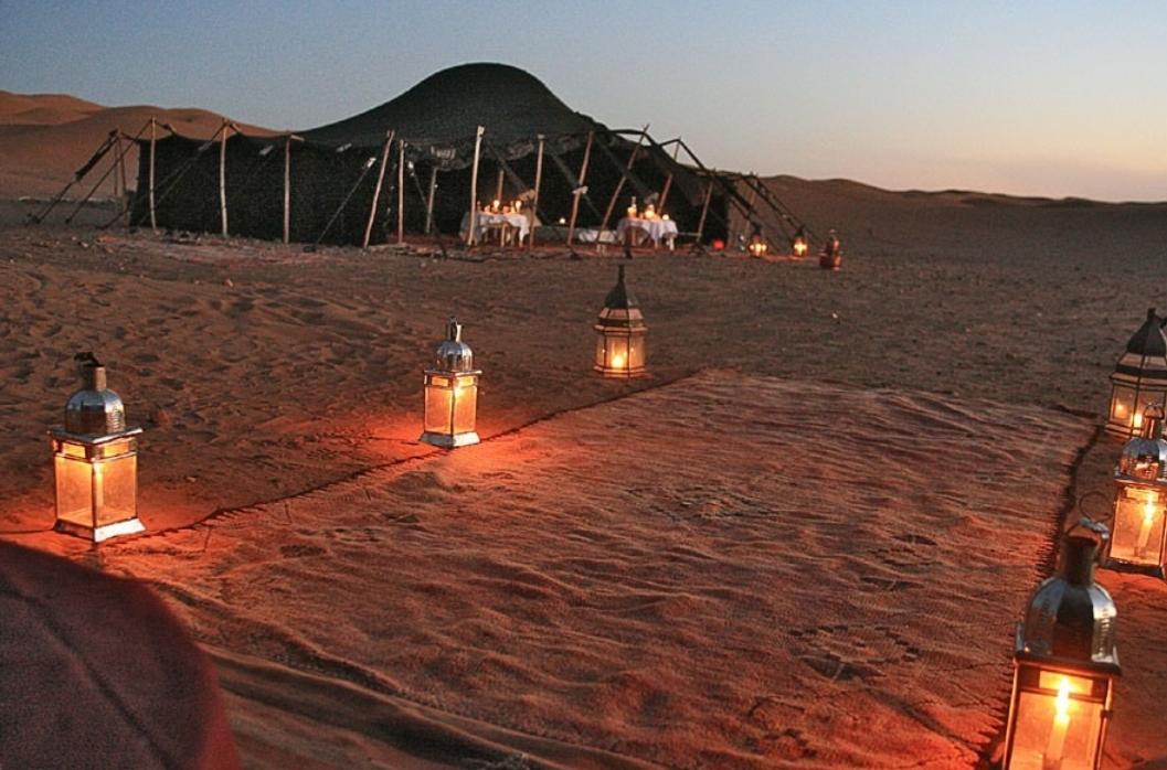 A traditional nomadic tent is set against a backdrop of sand dunes at dusk, surrounded by soft lighting from lanterns. A patterned rug is placed on the ground, leading to the tent entrance, where a communal space is visible for gathering.