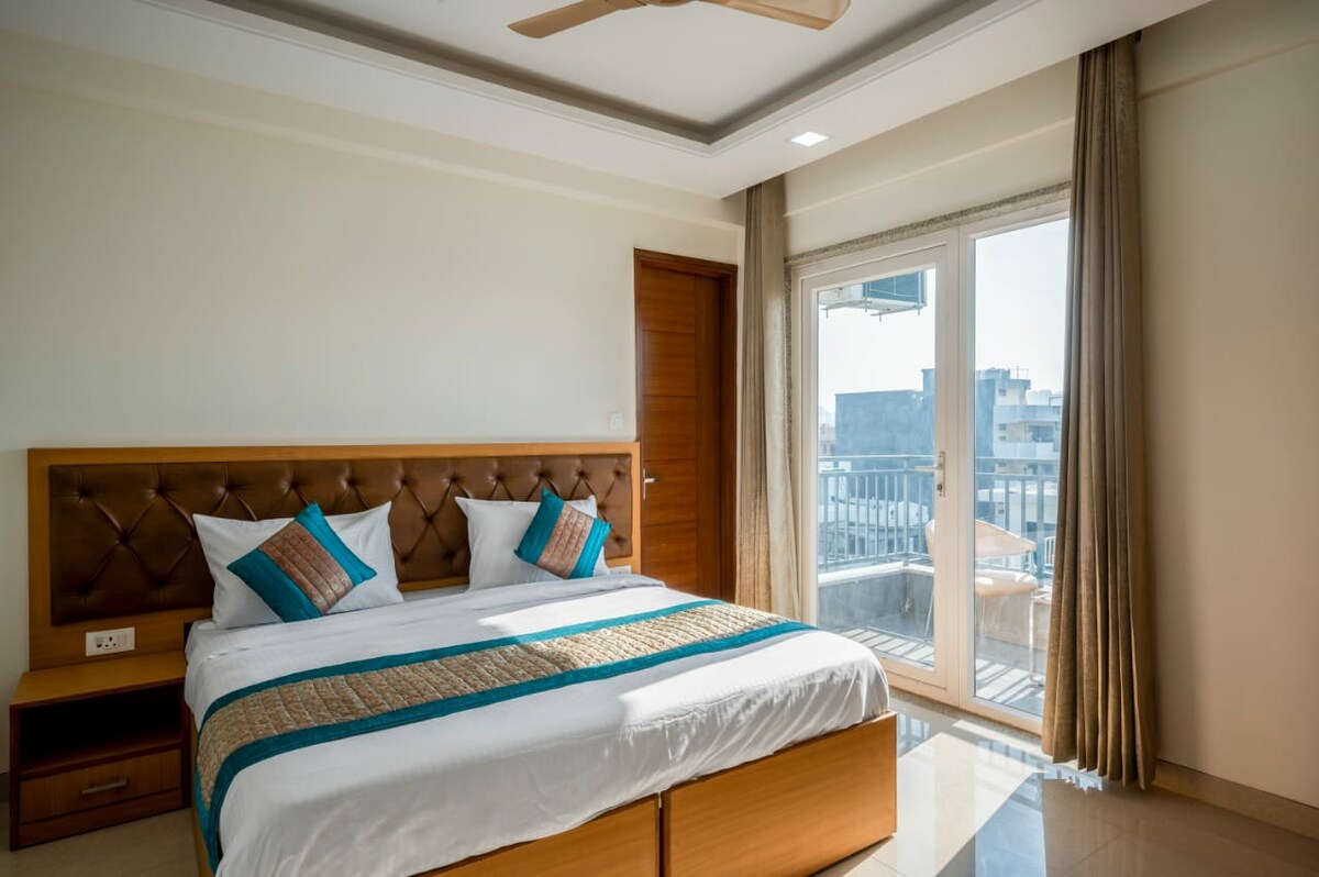 A well-appointed bedroom features a spacious bed with decorative cushions. Natural light enters through large glass doors that lead to a balcony, while soft curtains enhance the bright atmosphere. A cozy bedside table is placed on one side, completing the comfortable setting.