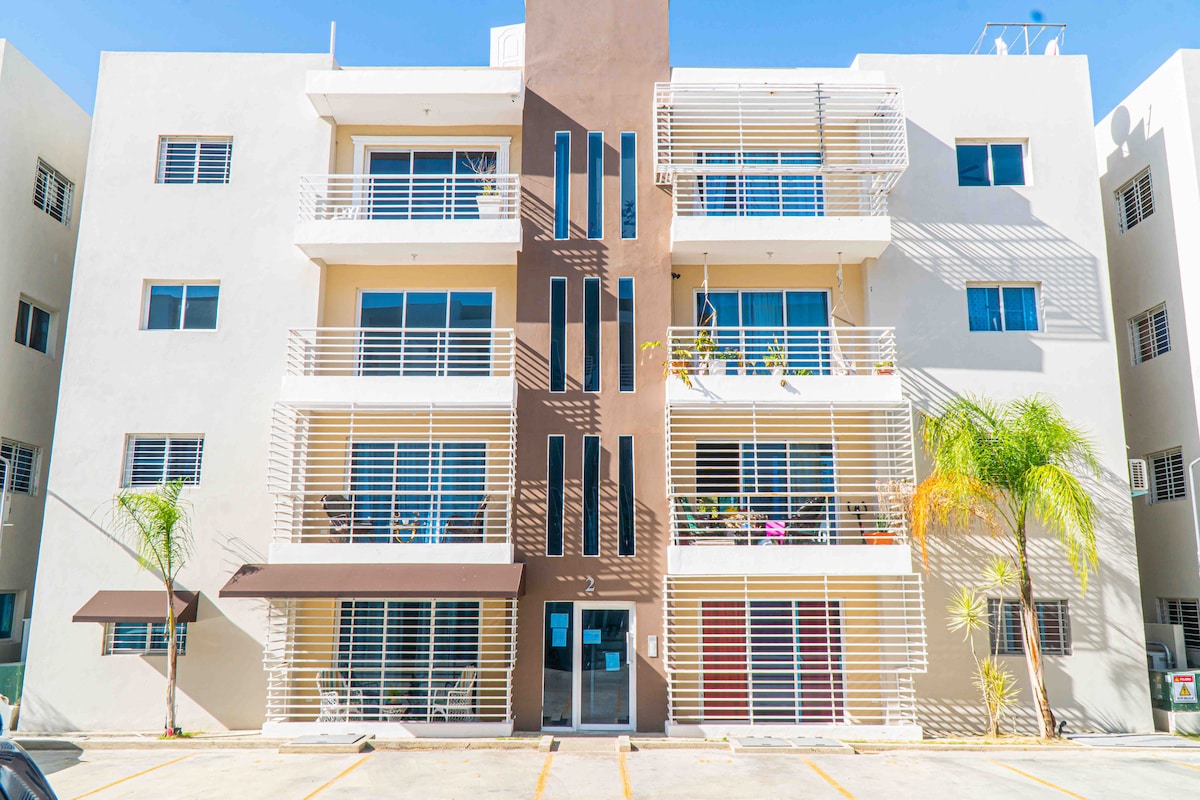 A modern three-story building features a symmetrical façade with large windows and balconies. Potted plants are visible on the balconies, complementing the clean lines of the exterior. A shaded entrance leads to the front door, while palm trees are situated on both sides.
