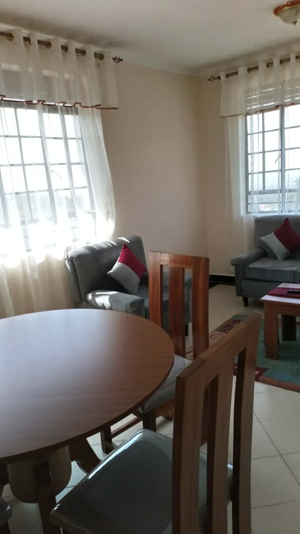A dining area features a round wooden table and four wooden chairs. Two comfortable chairs are positioned beside the table, near large windows that allow natural light to fill the space. Soft curtains frame the windows, creating a bright and welcoming ambiance.