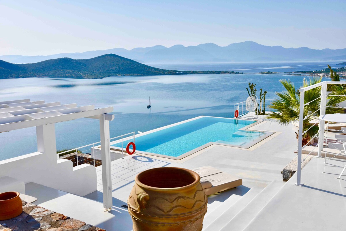 A panoramic view captures the inviting infinity pool overlooking the bay, framed by the tranquil waters and distant mountains. A terracotta pot sits prominently, adding a touch of character to the modern design. Sun loungers are available nearby for comfort and relaxation.