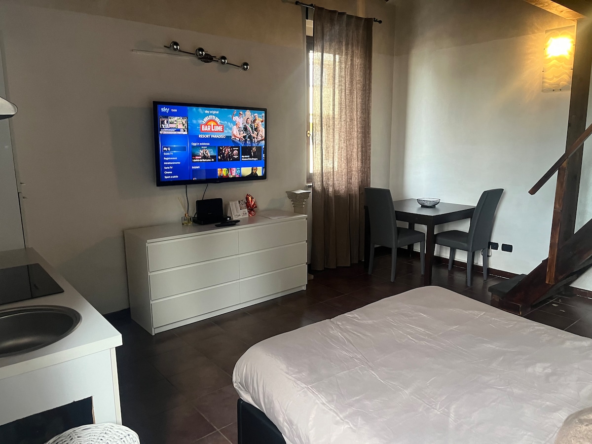 A modern living space features a spacious bed positioned in front of a large flat-screen TV mounted on the wall. A small dining table with two chairs sits nearby, accompanied by a kitchenette with a sink. Natural light enters through a window dressed with light curtains.