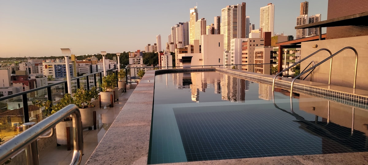 A rooftop pool presents a sleek design, surrounded by a modern deck. The tranquil water reflects the skyline of tall buildings, with evening light enhancing the urban view. Seating areas are visible in the background, suggesting a space for relaxation and enjoyment.