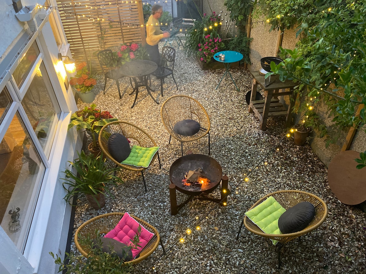 A charming courtyard is surrounded by greenery and twinkling fairy lights, suggesting a cozy atmosphere. Several wicker chairs with colorful cushions are positioned around a fire pit. Potted plants and a small table enhance the inviting outdoor space.