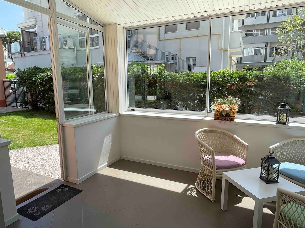 A bright veranda is shown with large windows offering views of the private garden. Two white wicker chairs are positioned beside a small table. Lanterns and a flower arrangement add to the serene ambiance, creating a welcoming space for relaxation.