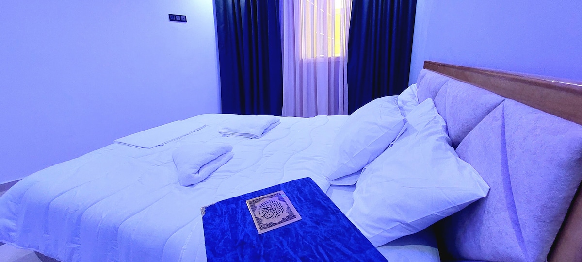 A spacious bed is dressed in crisp white linens, accompanied by neatly arranged pillows. A decorative blue cloth is placed at the foot of the bed. Soft light filters through sheer curtains, creating a gentle ambiance in the room.