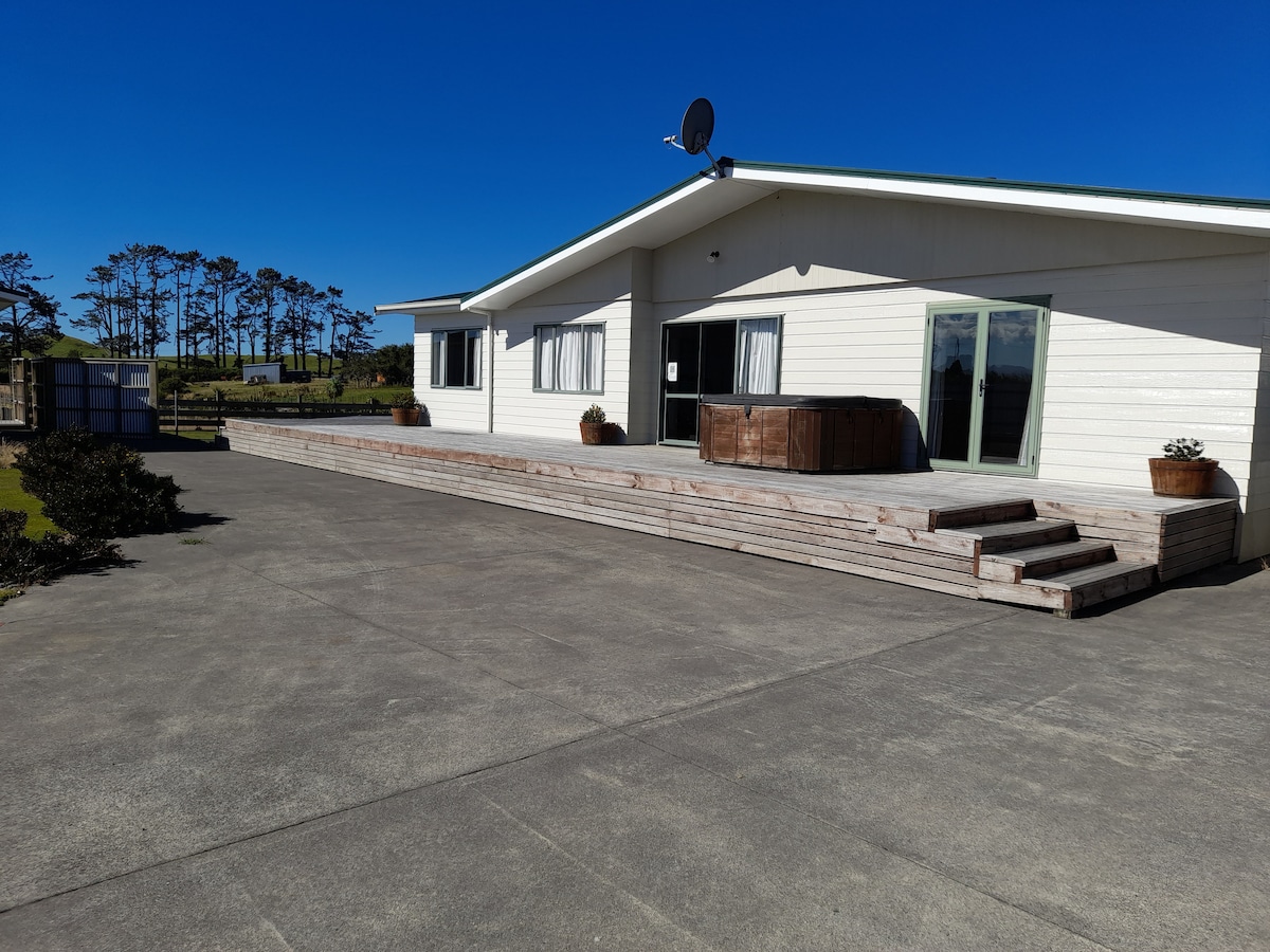 A spacious deck extends from the white-clad house, designed for outdoor relaxation. Large glass doors provide access to the interior, while planters add greenery. A hot tub is positioned on the deck, offering views of the surrounding landscape under clear blue skies.