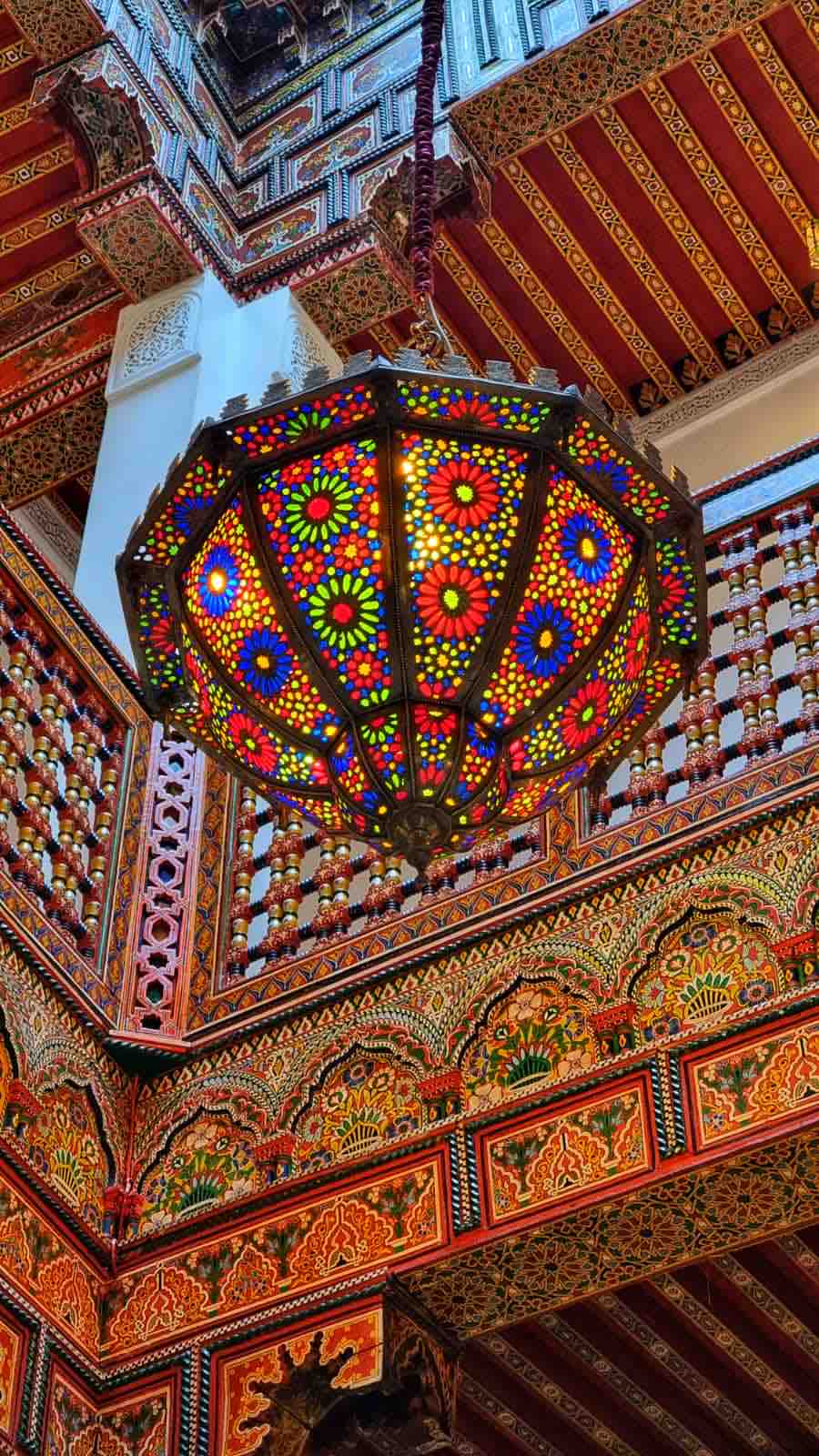 A vibrant multicolored lantern hangs from the ceiling, showcasing intricate floral patterns in red, blue, and yellow tones. Surrounding elements of traditional woodwork and ornate tile patterns enhance the rich cultural aesthetic of the space.