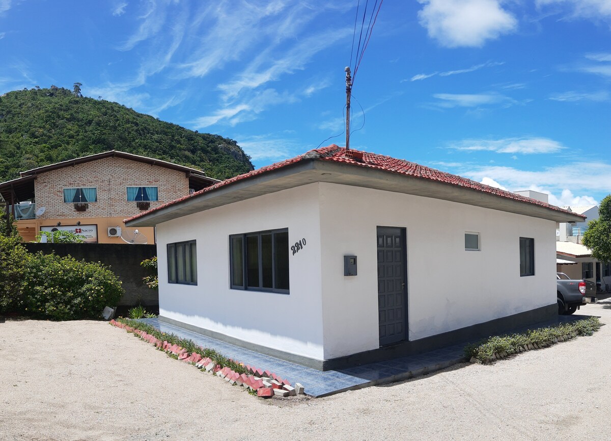 A simple, single-story house is presented with a flat roof and brick exterior. Large windows allow natural light to enter. A gravel pathway lined with plants leads to the entrance, while the structure is set against a backdrop of green hills and blue skies.