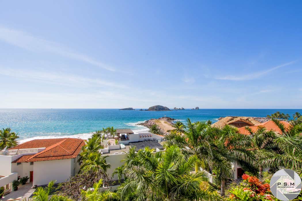 A panoramic ocean view is presented, featuring gentle waves lapping the shore. Lush palm trees are visible in the foreground, framing a serene beach scene. The distant coastline is marked by rocky outcrops and vibrant greenery, all set against a clear blue sky.