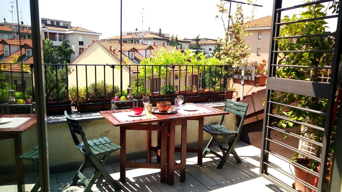 A terrace features a wooden table set for two, surrounded by greenery in pots. Two folding chairs are positioned alongside the table. The sunlit area provides a view of nearby buildings and rooftops, enhancing the space's inviting atmosphere.