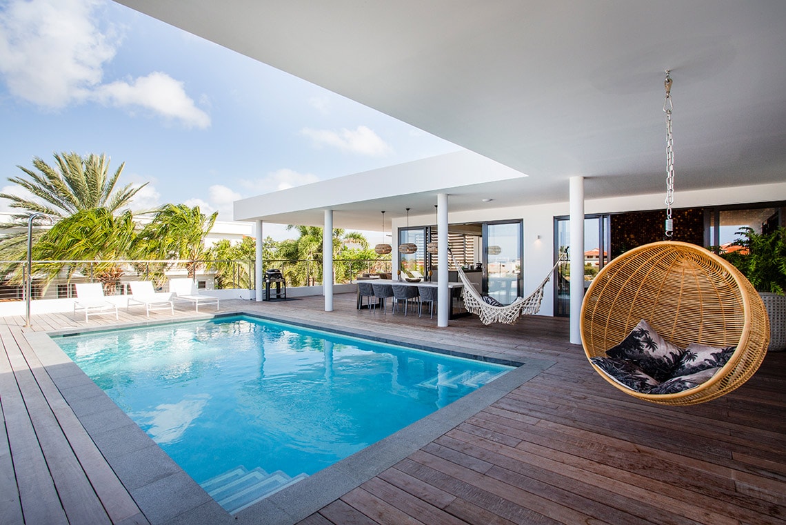 A sunlit patio features a serene swimming pool, surrounded by a wooden deck and comfortable lounge chairs. A hanging chair provides a cozy spot for relaxation. Tropical plants add greenery, while outdoor dining furniture is positioned under the covered area adjacent to the pool.