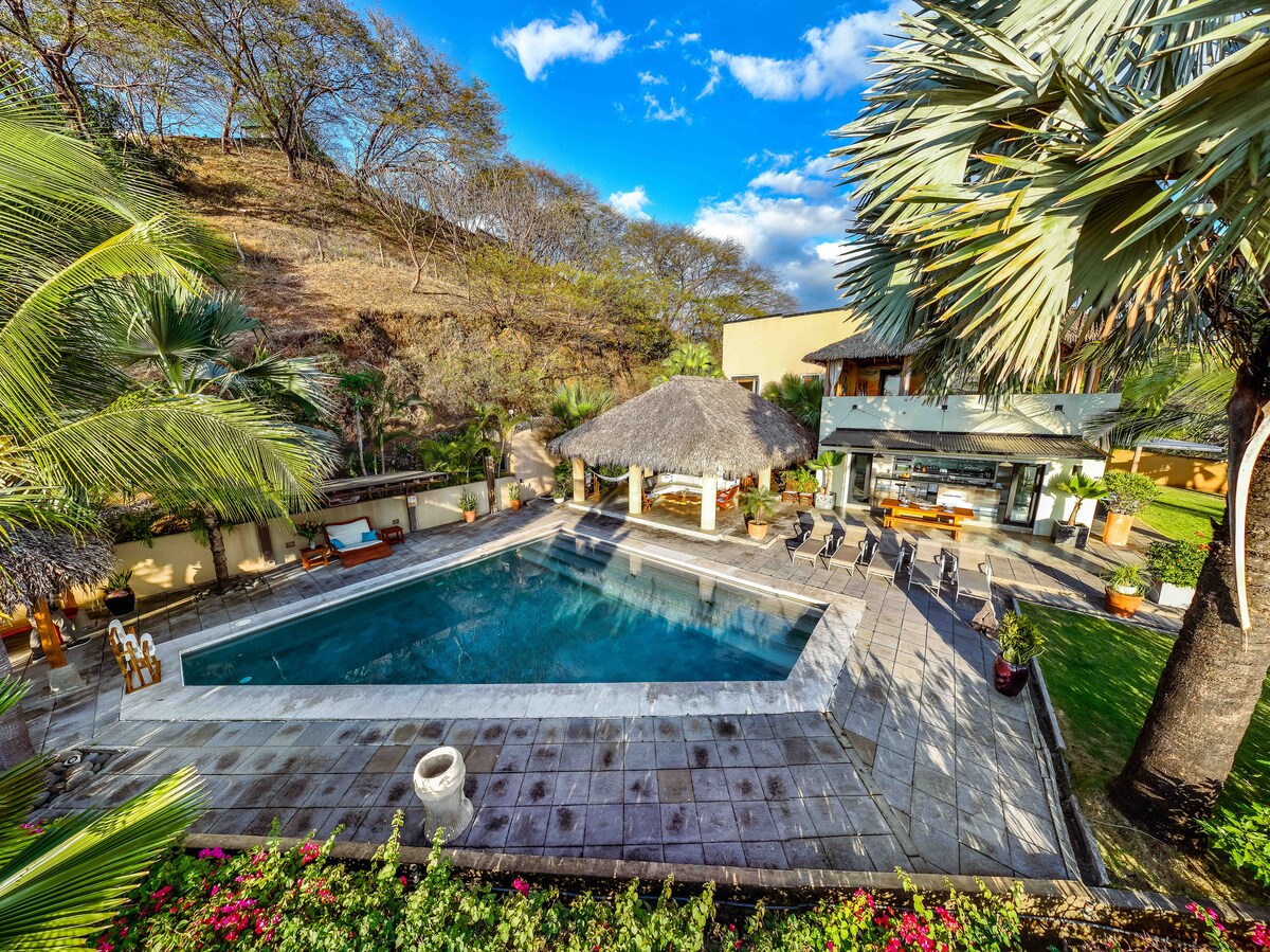 An inviting outdoor area features a spacious swimming pool surrounded by landscaped gardens. A palapa-style roof provides shade over a relaxation area. Comfortable seating is arranged nearby, with tables positioned for dining. Lush greenery and palm trees enhance the tranquil atmosphere.