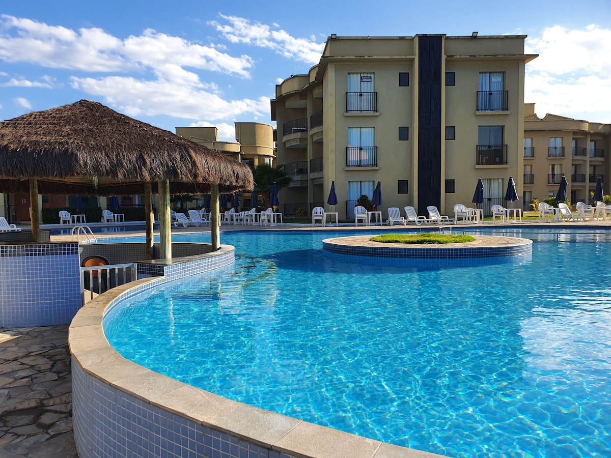 A large swimming pool, designed with a circular island in the center, features crystal-clear blue water. Surrounding the pool, a thatched-roof gazebo is visible, along with lounge chairs arranged neatly on the pool deck, set against a backdrop of the condominium building.