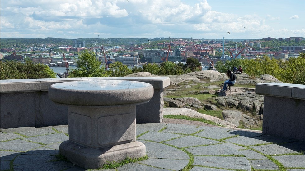 A stone observation table is centered in the foreground, positioned on a natural rock platform. Lush greenery surrounds the area, with cityscape views stretching into the distance under a partly cloudy sky. People can be seen engaging with the landscape, enjoying the serene environment.