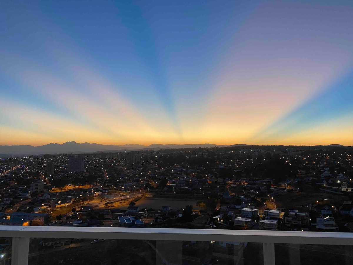 A panoramic sunset view depicts a gradient of colors across the sky, transitioning from deep blue to golden yellow. Silhouettes of distant mountains are visible on the horizon, while city lights twinkle below, creating a serene and expansive backdrop.