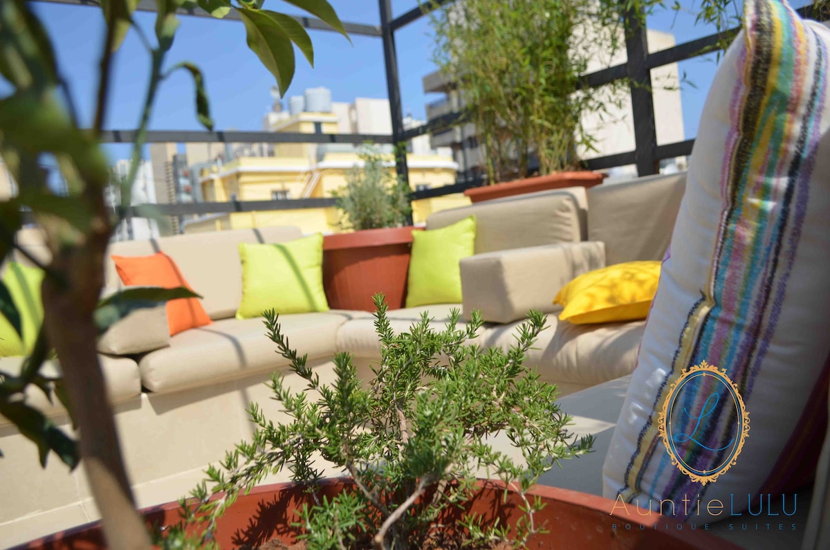 A rooftop lounge area is visible, featuring a light-colored sectional sofa adorned with colorful cushions. Potted plants are strategically placed, enhancing the space. Clear skies provide bright natural light, creating an inviting environment for relaxation.