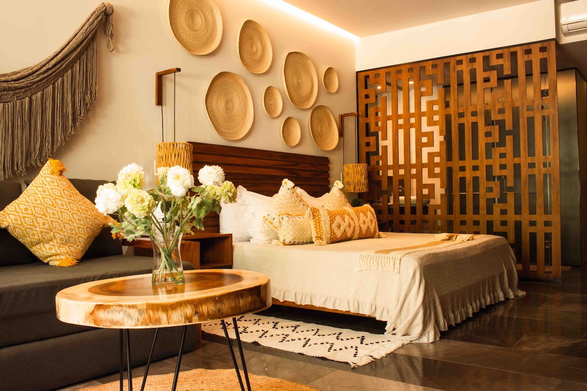 A well-furnished studio features a warm color palette, highlighted with woven wall decor and decorative cushions. A wooden partition adds visual interest, while a vase of fresh flowers sits atop a round wooden table, contributing to the inviting ambiance.
