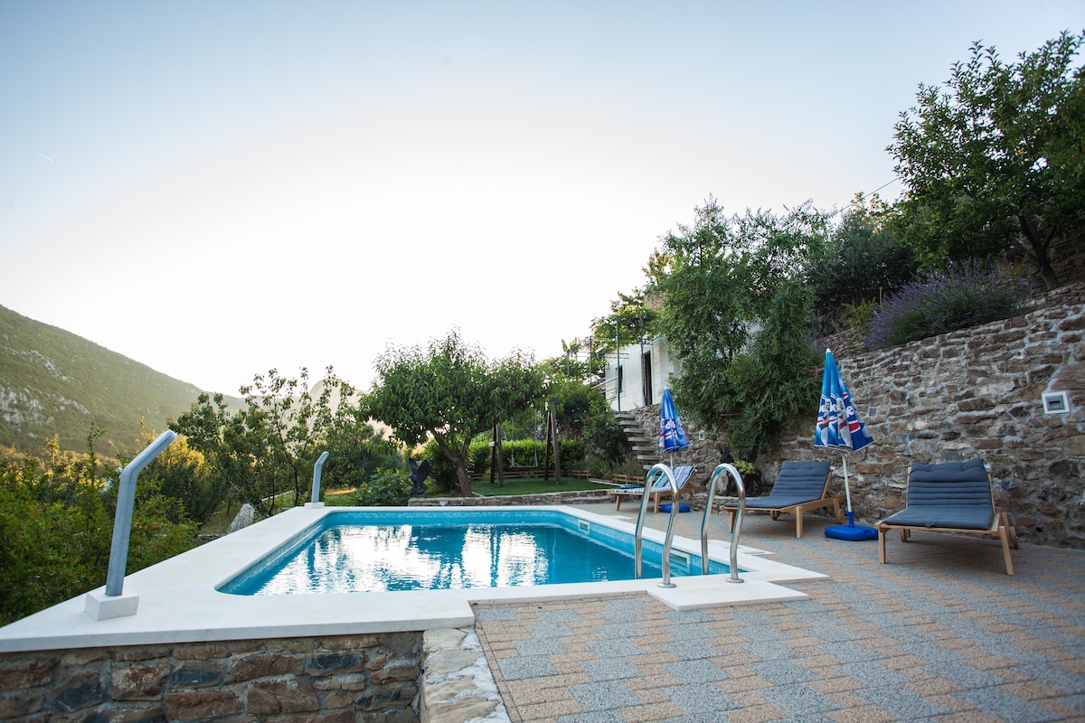 A heated swimming pool is surrounded by stone paving and lush greenery. Two blue lounge chairs and umbrellas sit nearby, with trees and plants framing the space. The backdrop features gentle hills, creating a serene countryside setting.