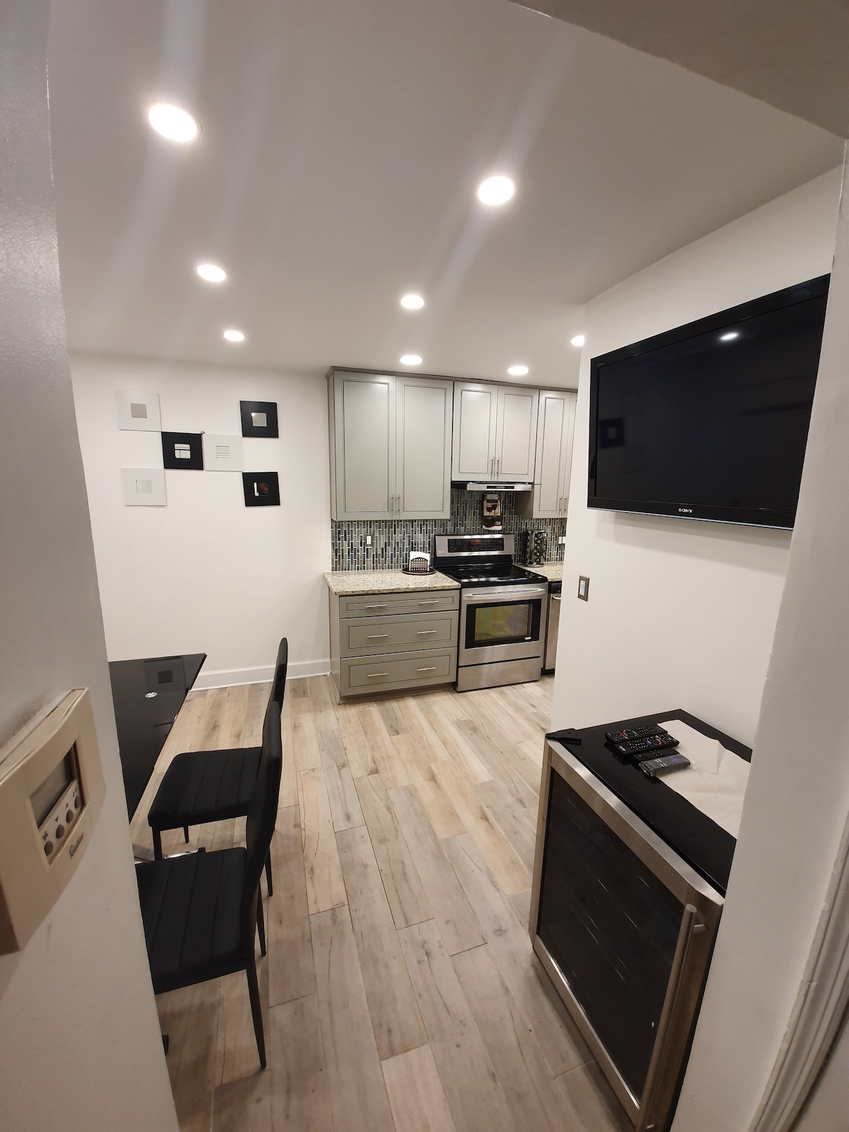 The modern kitchen is equipped with sleek cabinetry and stainless-steel appliances, including an oven and refrigerator. A dining table with chairs is positioned nearby, while ambient lighting enhances the space. A wall-mounted television is visible, adding to the contemporary feel.