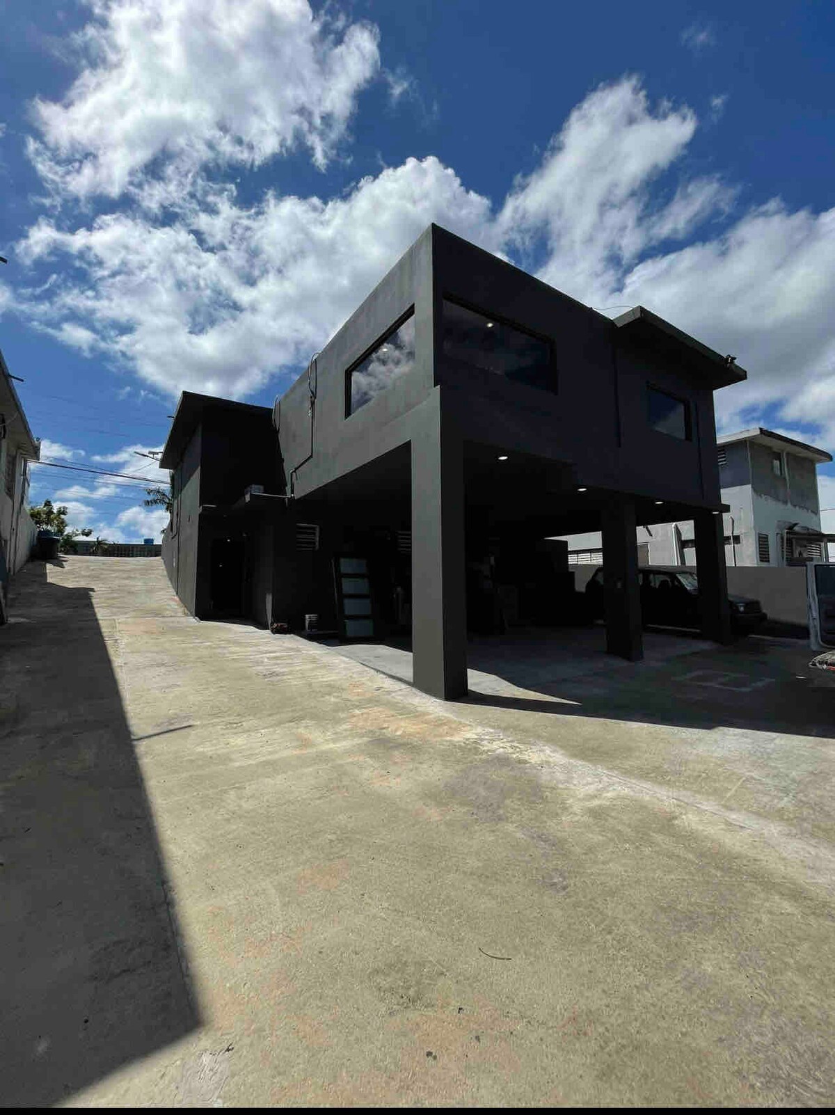 A modern structure stands prominently against a bright blue sky, featuring a sleek black exterior. The building is elevated with a spacious driveway leading up to it, showcasing large windows that allow natural light to flow indoors.