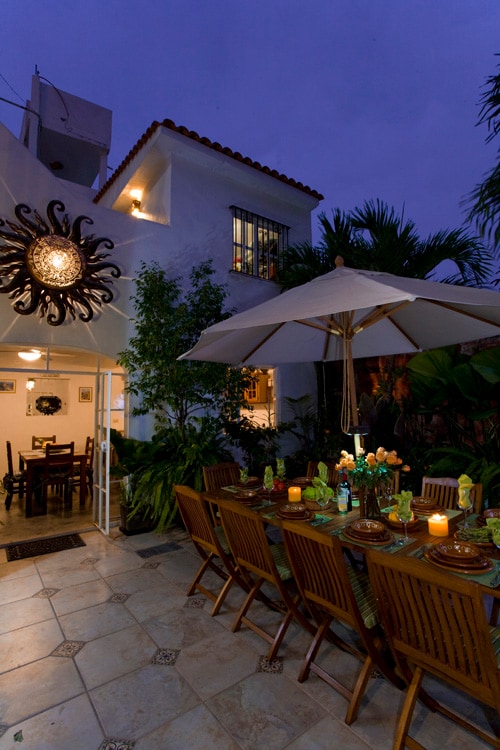 An outdoor dining area is illuminated at dusk, featuring a large table with wooden chairs under a white umbrella. Lush greenery surrounds the space, with candles and decorative items adding a touch of charm. A rustic wall sconce provides additional warm lighting.
