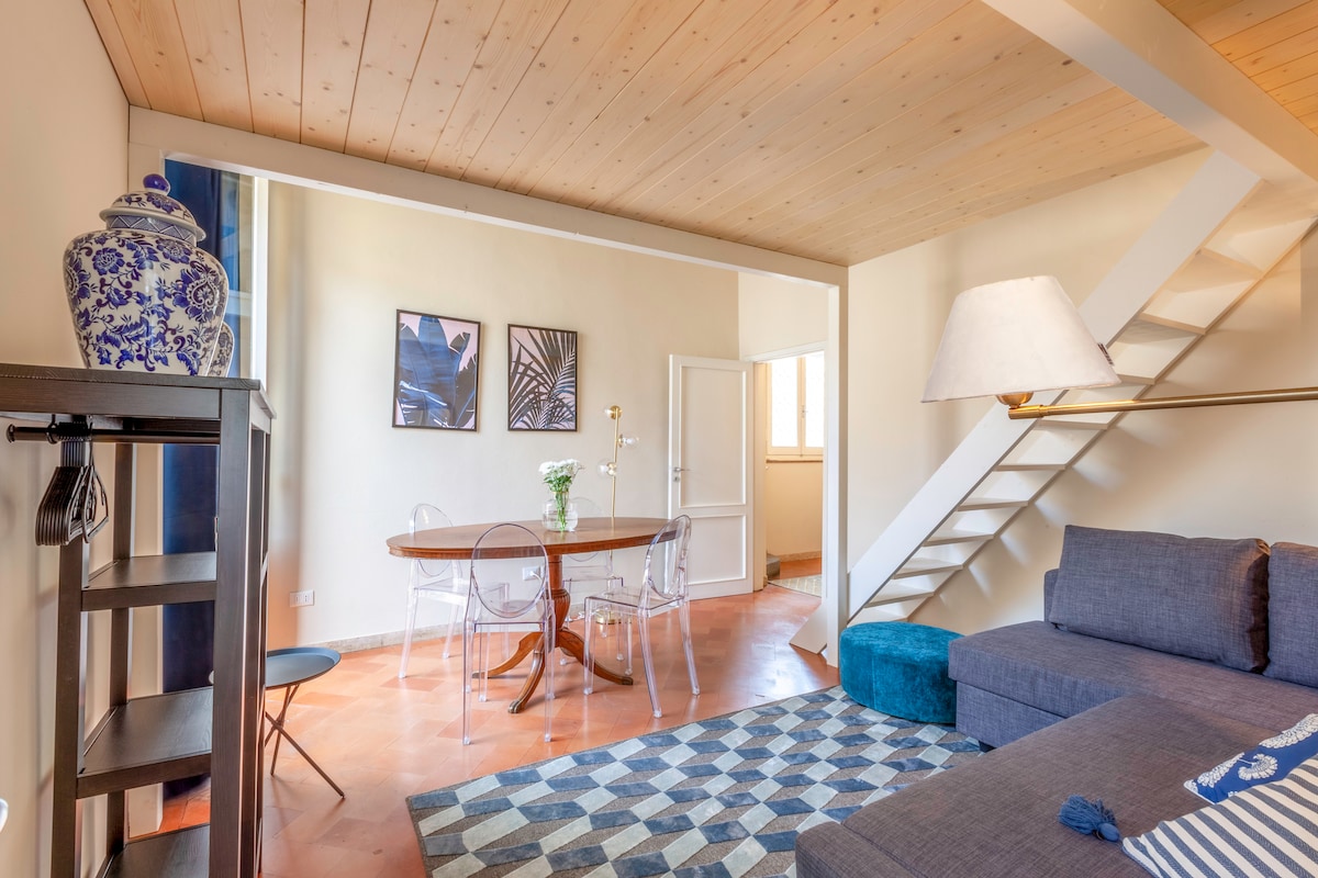 A light-filled living area features a modern, spacious gray sofa and an elegant dining table set for two. Clear views of the staircase lead to the upper loft, while decorative artwork adorns the walls. A blue pouf adds a touch of color to the neutral palette.
