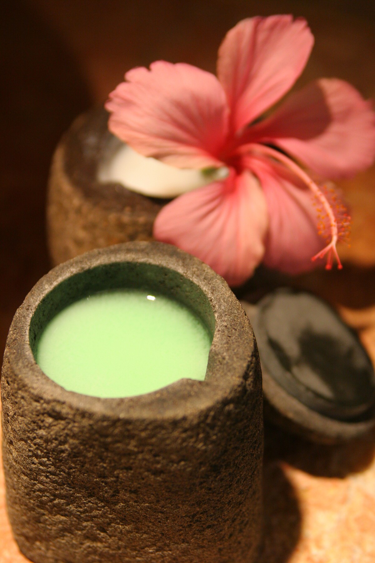 Two stone containers are placed beside a vibrant pink hibiscus flower. One container is slightly open, revealing a bright green liquid inside. The surface beneath features a warm, natural texture, enhancing the overall aesthetic.