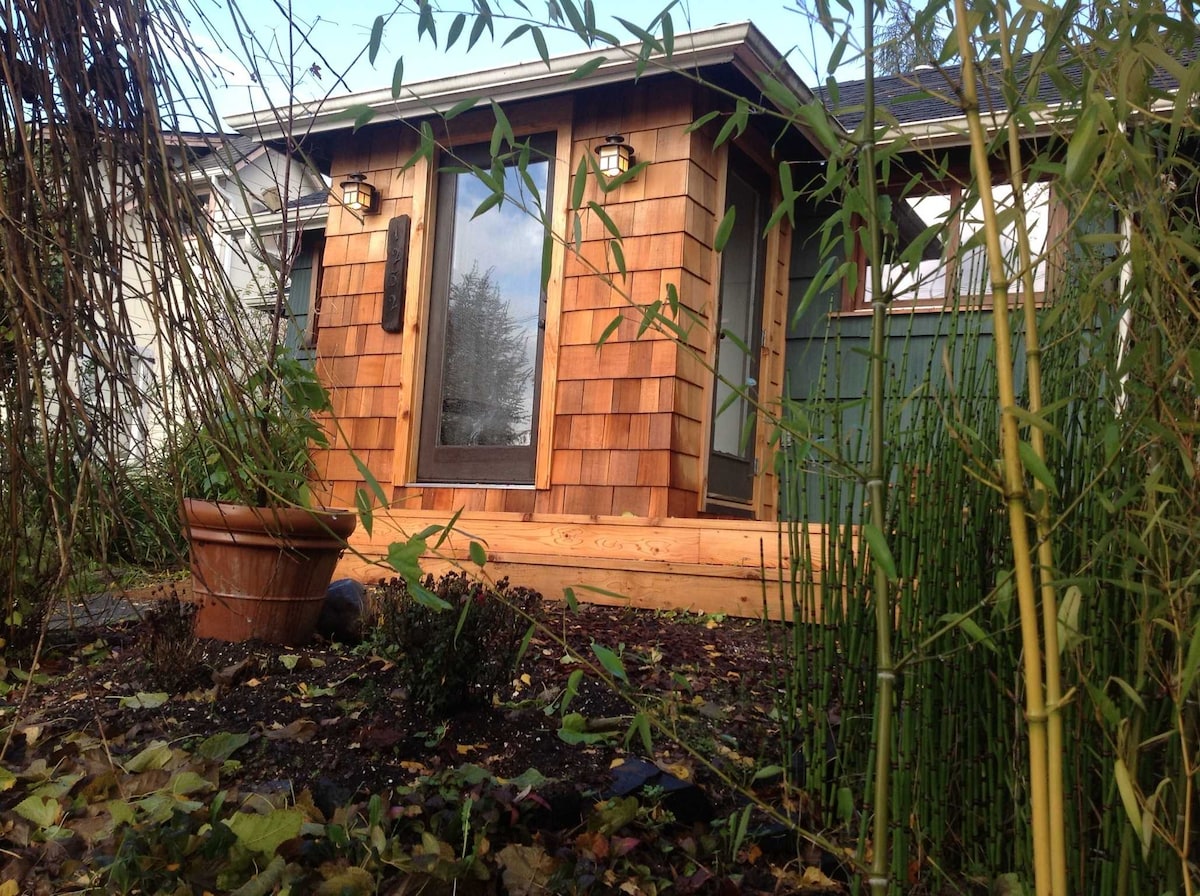 The exterior of the Bungalow is framed by lush greenery and bamboo, highlighting the wooden shingle facade and large glass doors. A flower pot is positioned near the entrance, which is set against a peaceful, natural backdrop.