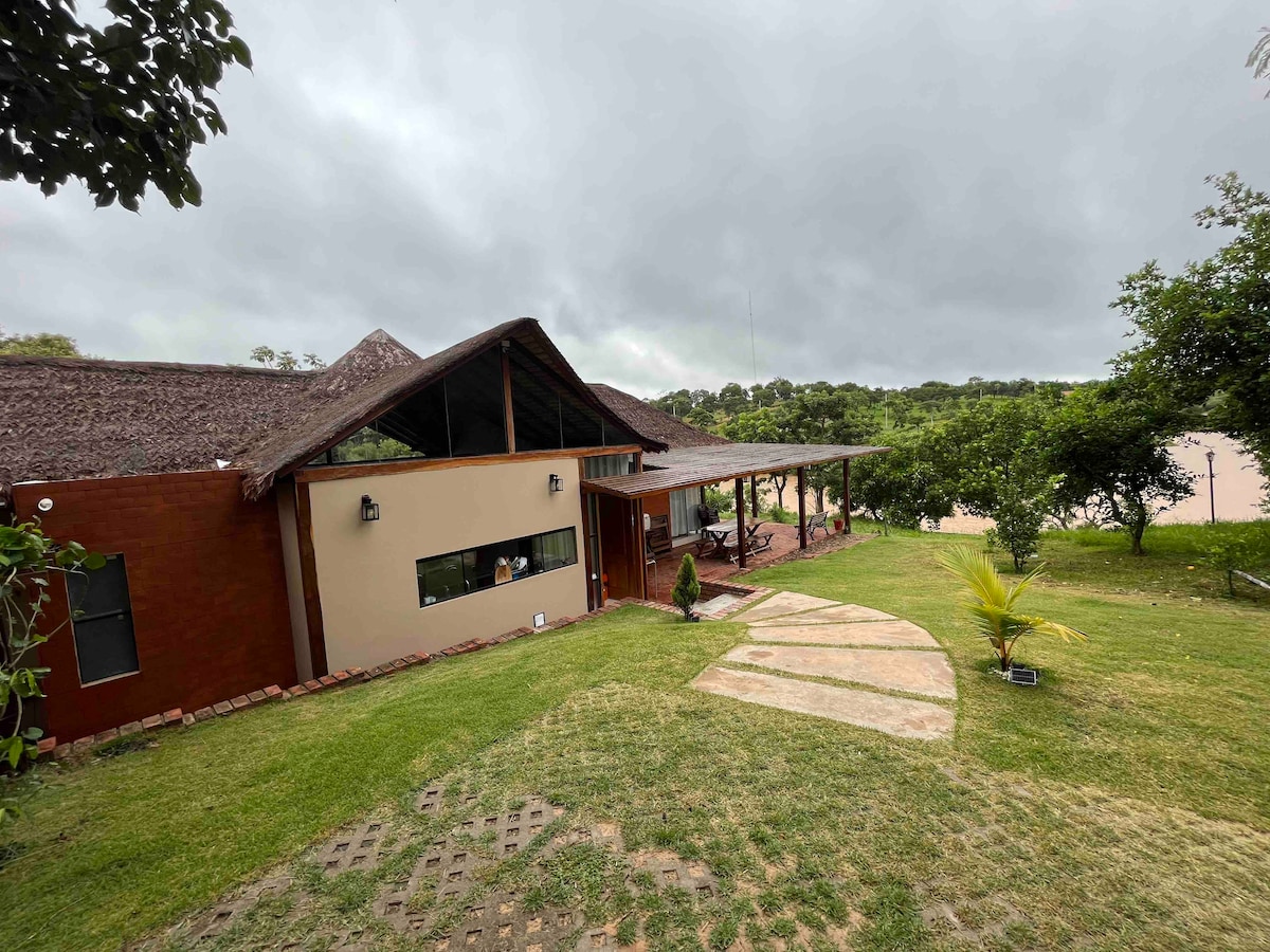 The cabin is set amidst greenery, featuring a thatched roof and large windows that allow natural light. A pathway leads up to the entrance, which is flanked by grass and small decorative plants. The surrounding area includes trees and an expansive view of the landscape.