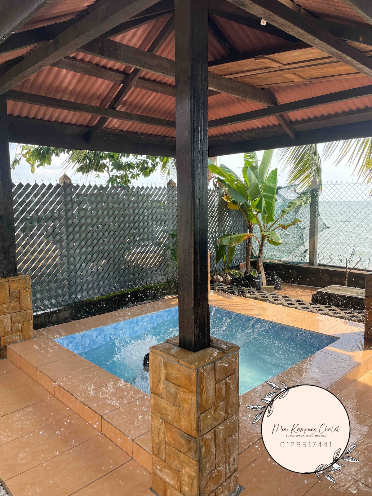 A covered gazebo area features a small pool surrounded by stone pillars and tiled flooring. Tropical plants are visible beyond the structure, with a fence providing a sense of privacy. Water can be seen gently splashing in the pool, creating a calming atmosphere.
