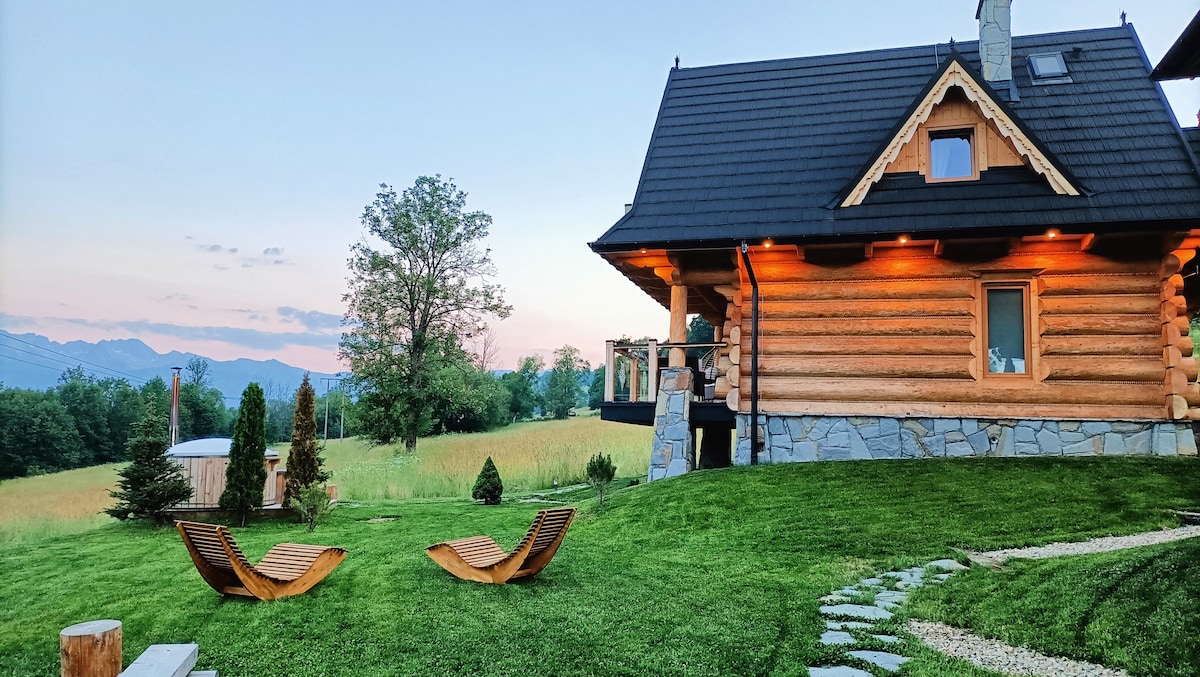 A log cabin stands surrounded by a lush green lawn, featuring wooden loungers for relaxation. In the background, rolling hills and distant mountain peaks are visible against a soft evening sky. The exterior is illuminated gently, enhancing the cabin's welcoming appearance.