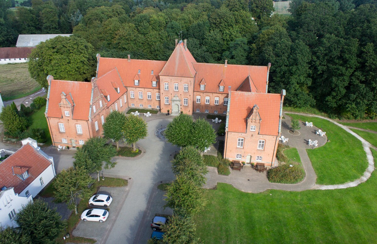 An aerial view of a historic building with a distinct architectural style featuring a red roof and ornate details. Surrounding green spaces are visible, along with pathways and several seating areas, inviting a sense of tranquility.