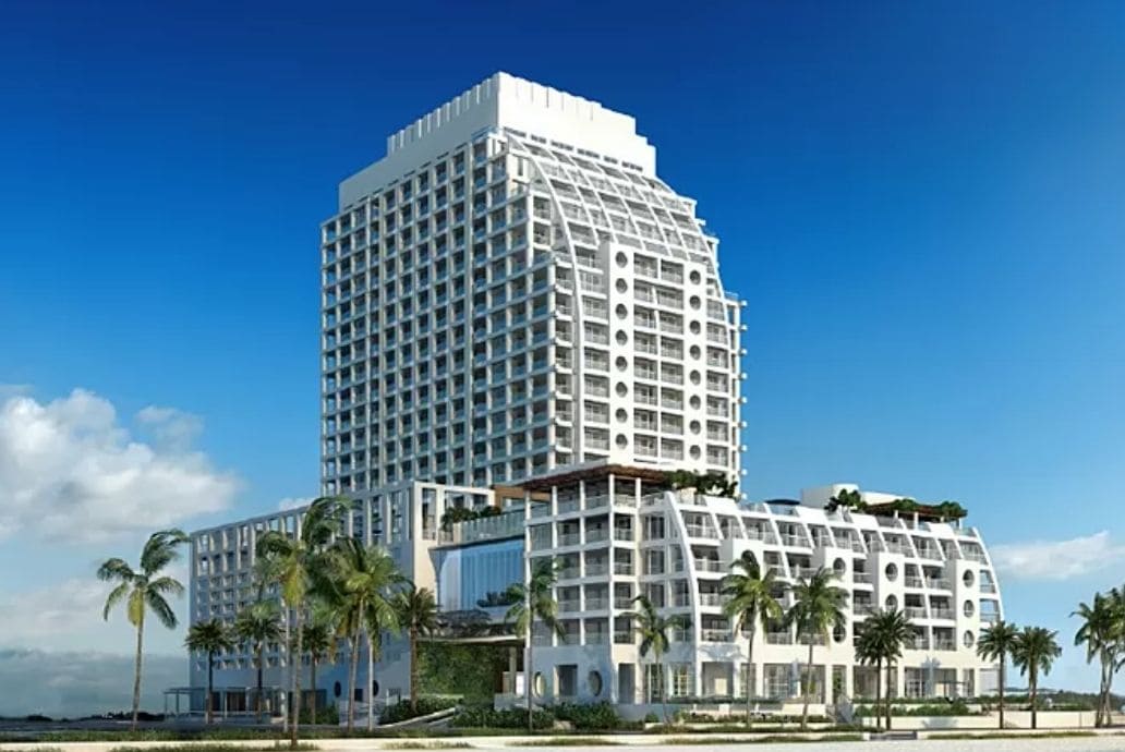 A modern beachfront hotel features a striking architectural design with multiple levels and balconies. Tall palm trees surround the property, and the bright blue sky enhances the inviting atmosphere of this luxurious setting near the ocean.