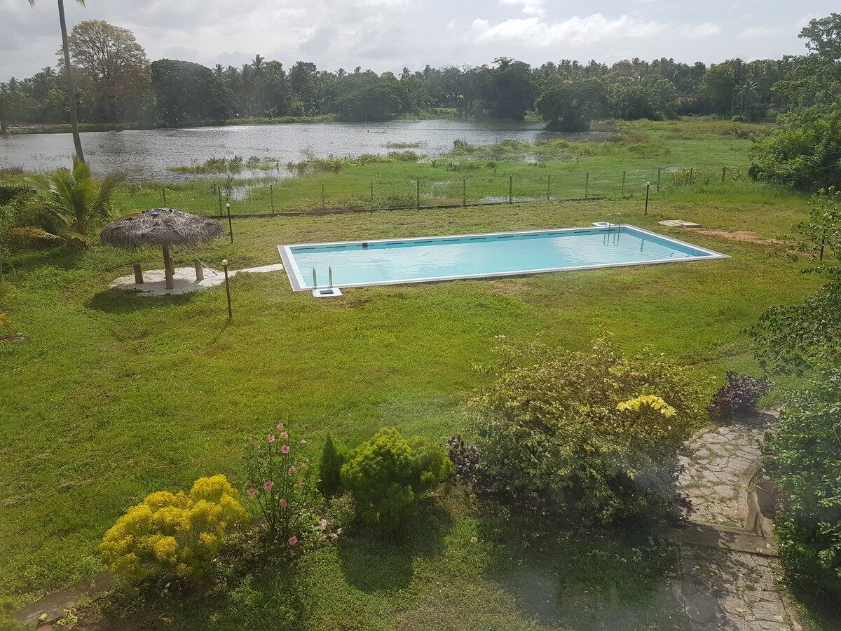 A clear view of a private pool surrounded by lush green grass is seen, with a thatched-roof gazebo positioned nearby. The calming expanse of a lake is visible in the background, framed by trees and gentle hills.