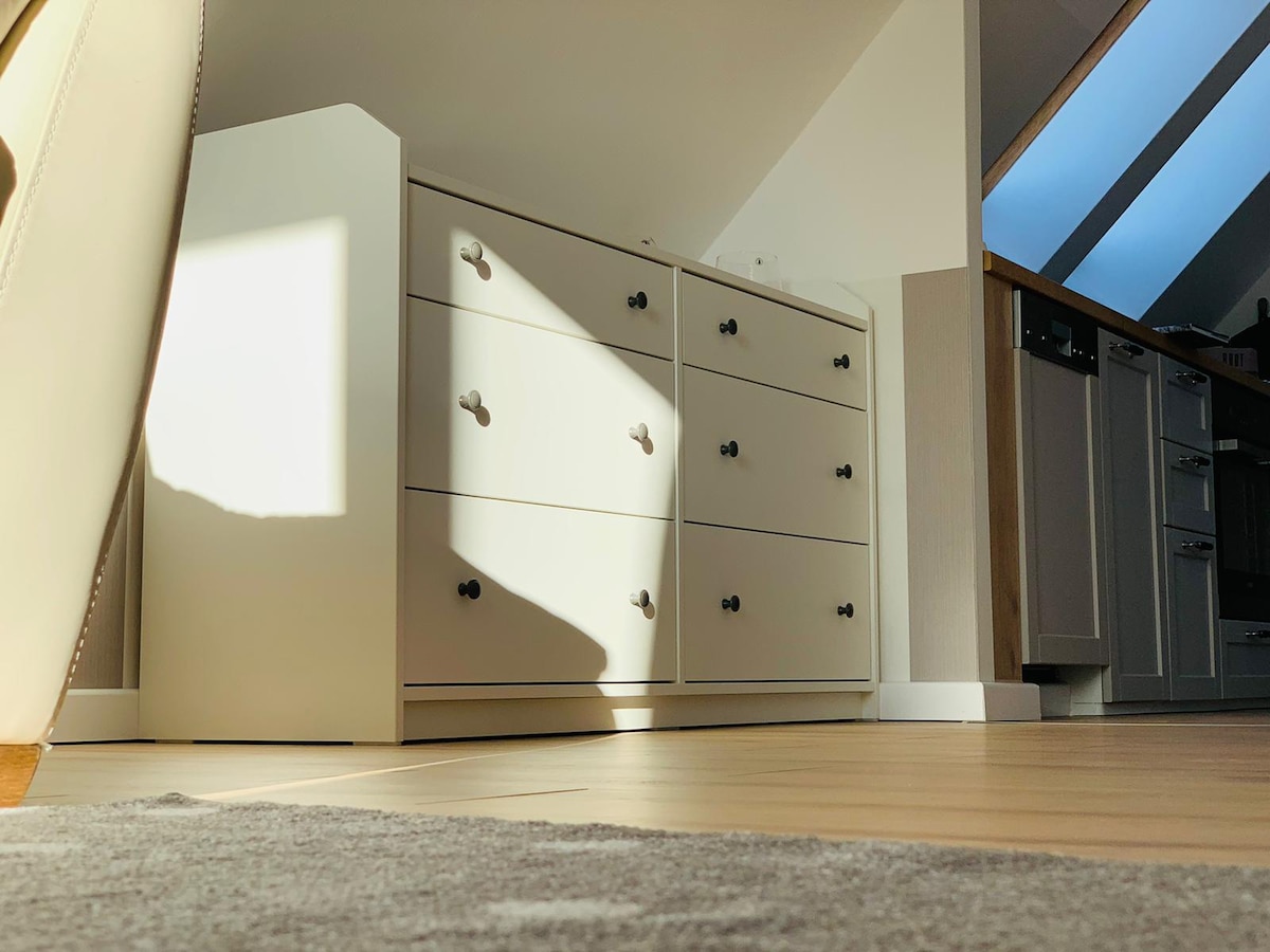 A white dresser with six drawers is positioned against a wall. Natural light streams in through skylights, casting soft shadows across the surface. The floor features light wooden planks, complemented by a neutral area rug in the foreground.